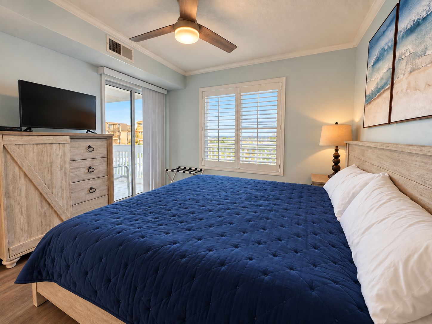 A bedroom with a bed, a television, a ceiling fan, a window with blinds, and artwork on the wall.