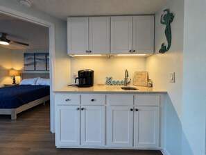 A compact bedroom with a bed and a nightstand, a kitchenette with a coffee maker, and a wall-mounted decorative piece. - A Place at the Beach III, Oceanfront Family-Friendly Resort (Atlantic Beach)