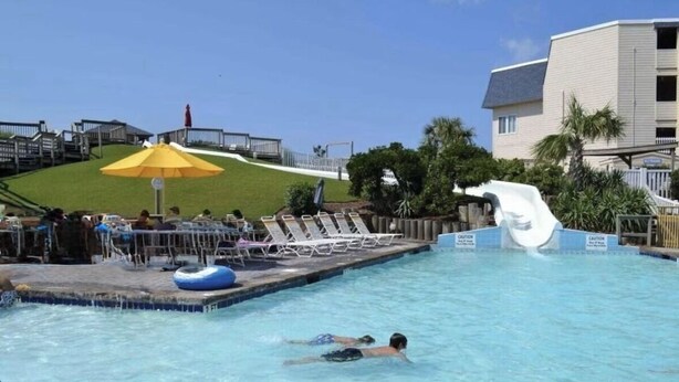 A swimming pool with a water slide, lounge chairs, and a grassy hill in the background.