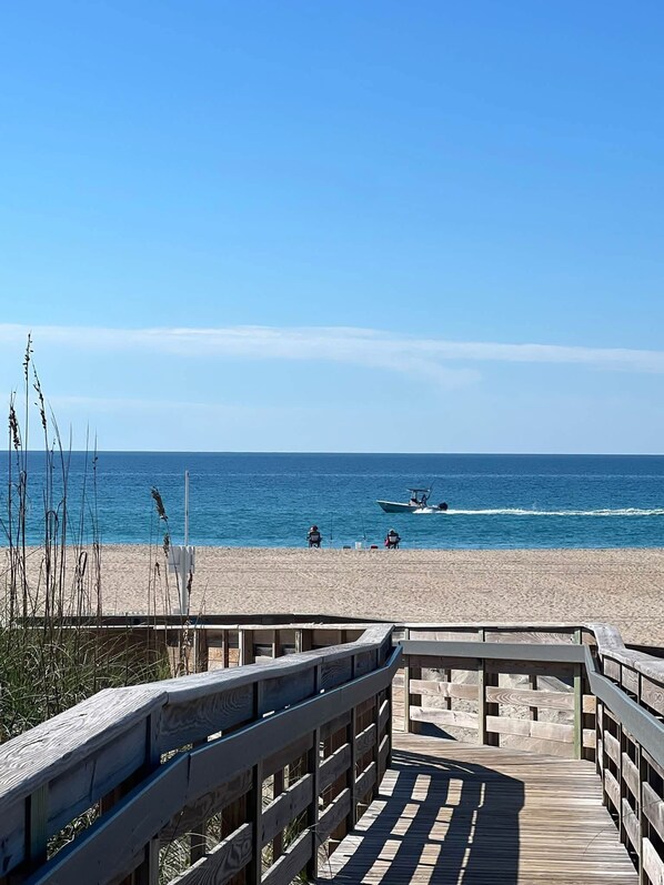 A wooden boardwalk leading to a beach with a clear view of the ocean and a boat in the distance.
