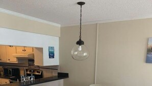 A dining area with a white table and chairs, a hanging light fixture, and a kitchen in the background.