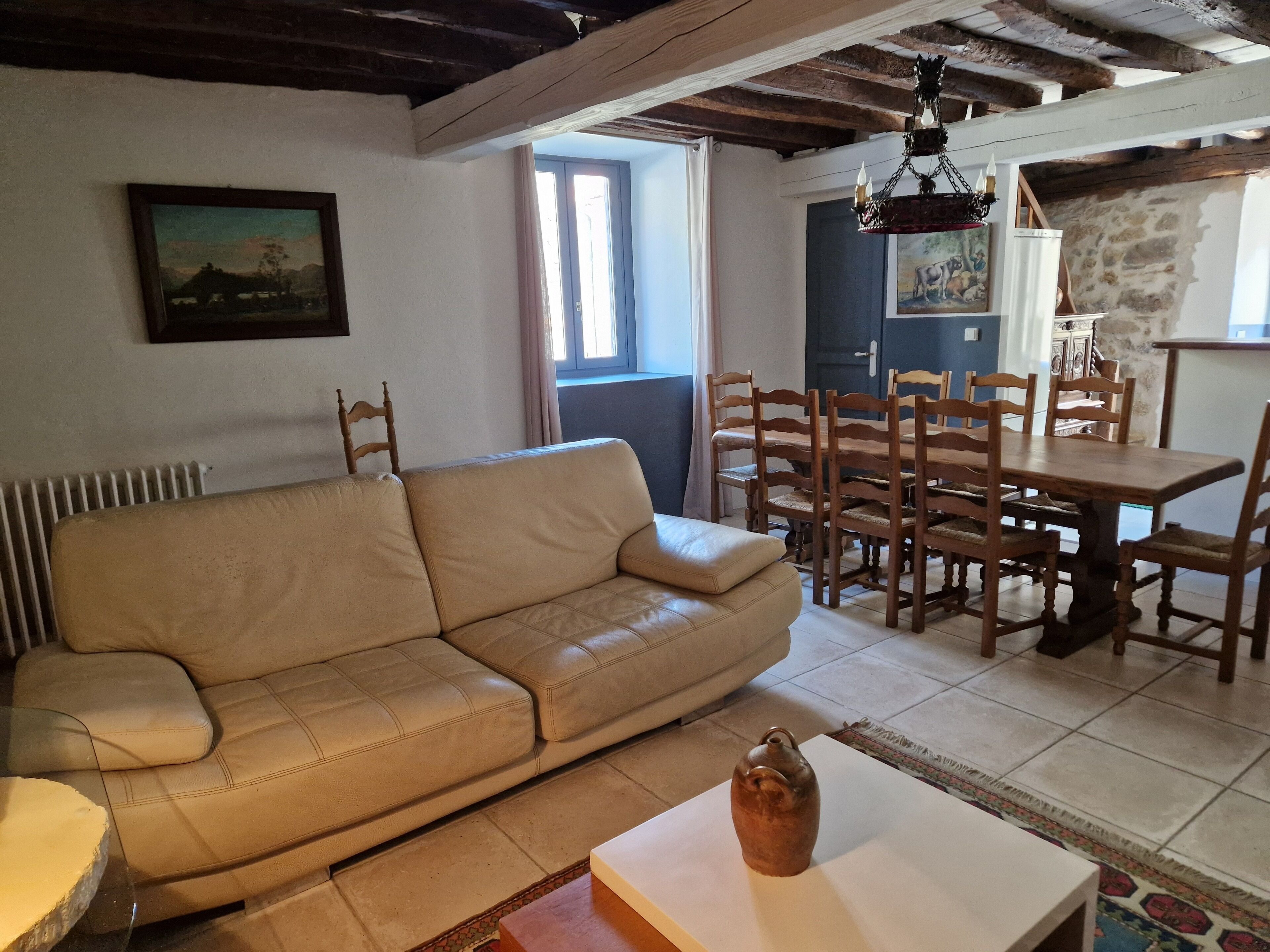 Een woonkamer met een beige bank, een houten eettafel met stoelen, een vaas op een tafel en een schilderij aan de muur.