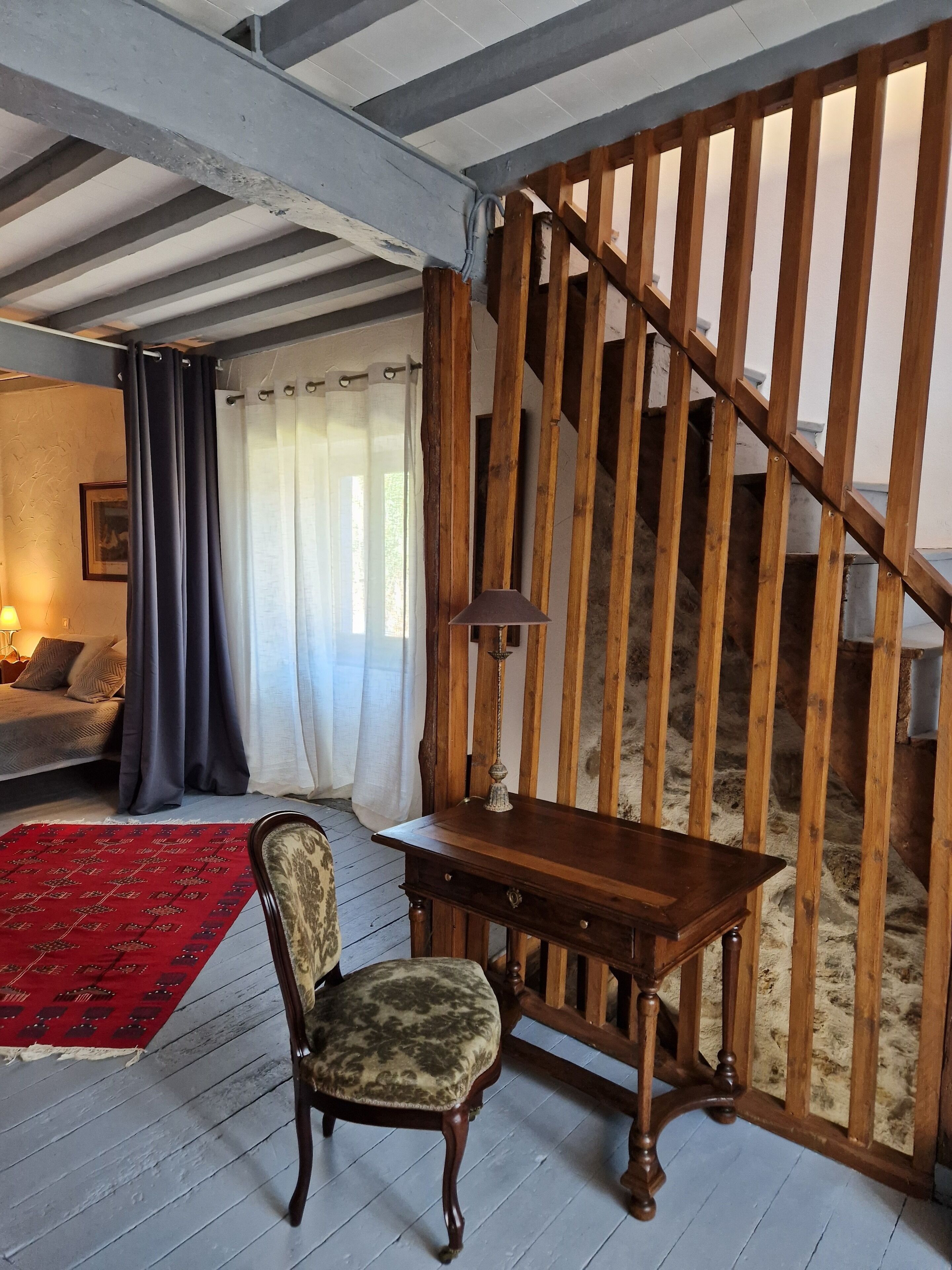 Ein Zimmer mit einer Holztreppe, einem Schreibtisch aus Holz, einem gemusterten Sessel, einem roten Teppich und einem Bett mit Lampe.