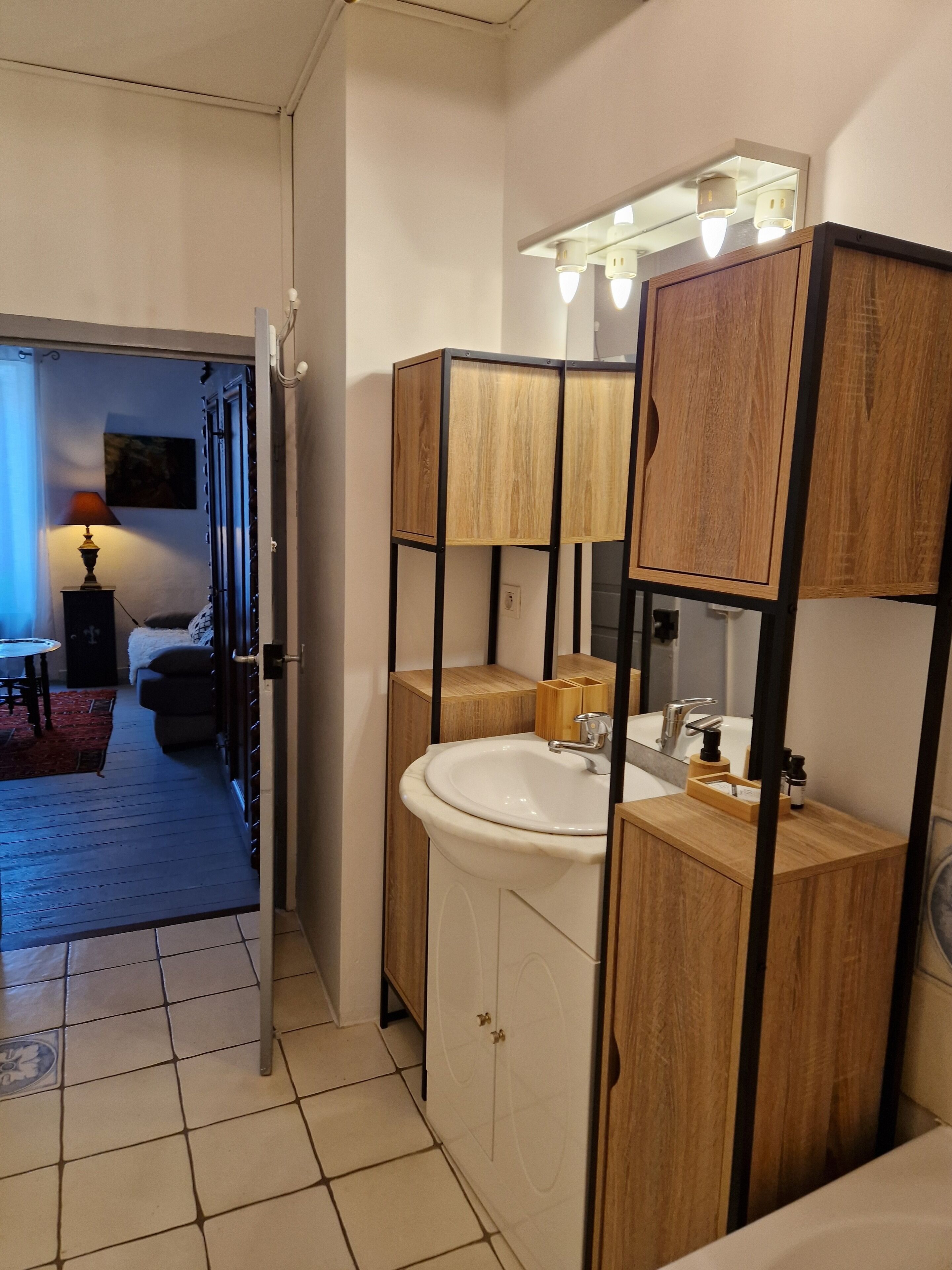 Een moderne badkamer met een houten wastafelmeubel, een wastafel en een spiegel. Er is een deur naar een slaapkamer met een bed en een nachtkastje.