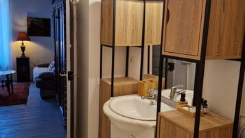 Een moderne badkamer met een houten wastafelmeubel, een wastafel en een spiegel. Er is een deur naar een slaapkamer met een bed en een nachtkastje.
