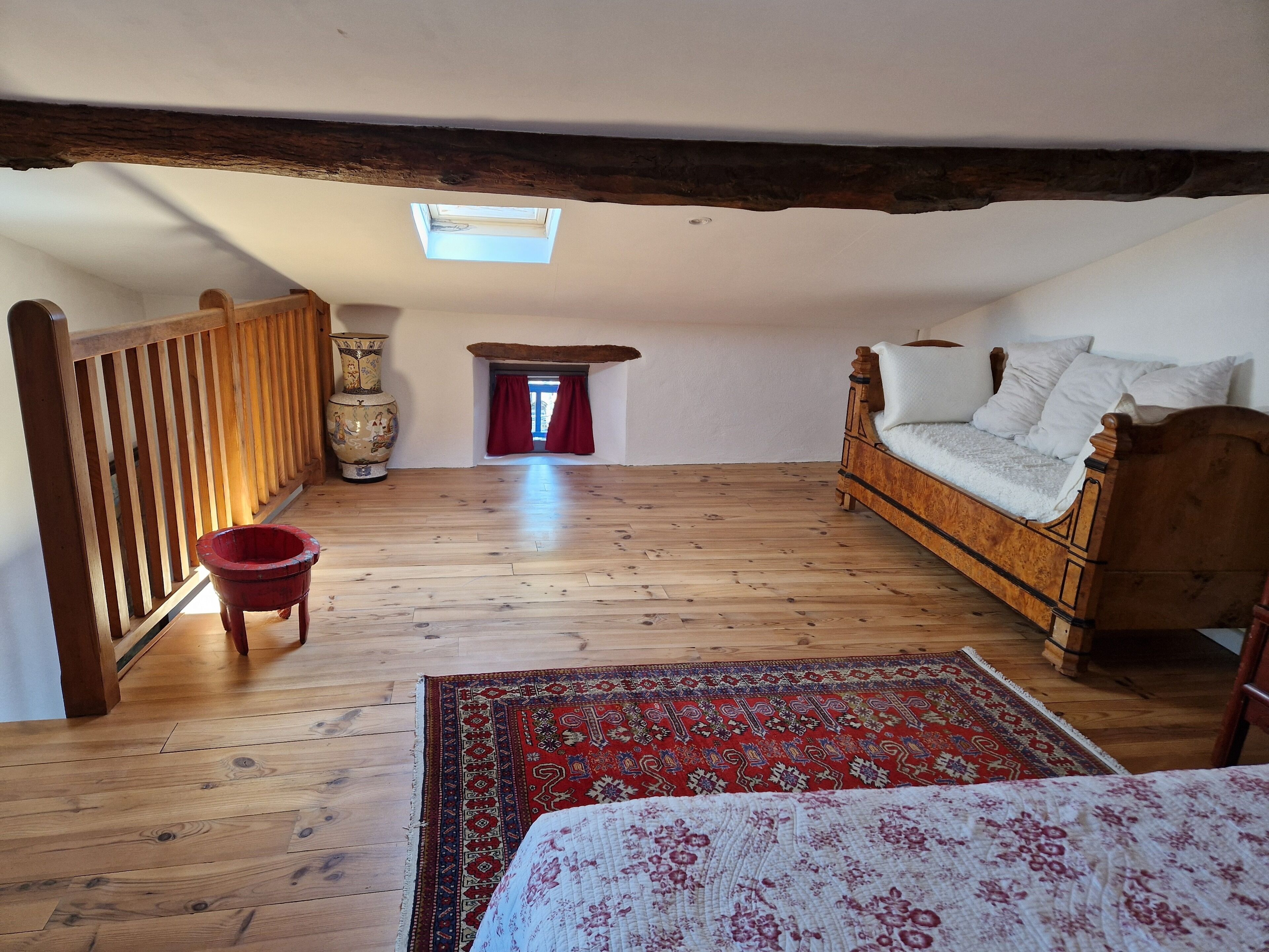 A room with a wooden floor, a bed with white bedding, a red rug, a wooden chair, a large vase, and a small window with red curtains.