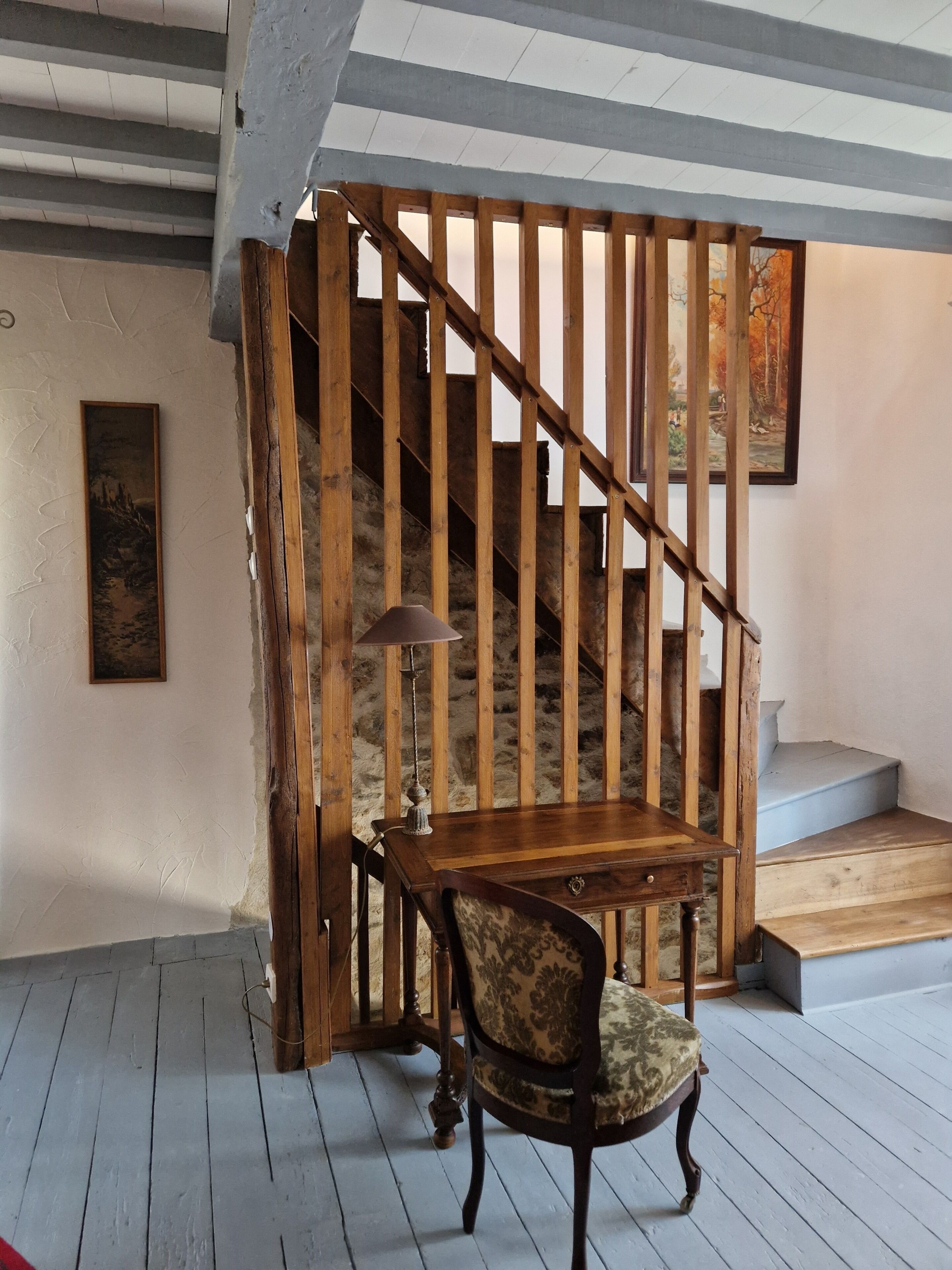 Una escalera con barandillas de madera, una lámpara y una silla en una habitación con pared texturizada y un cuadro.