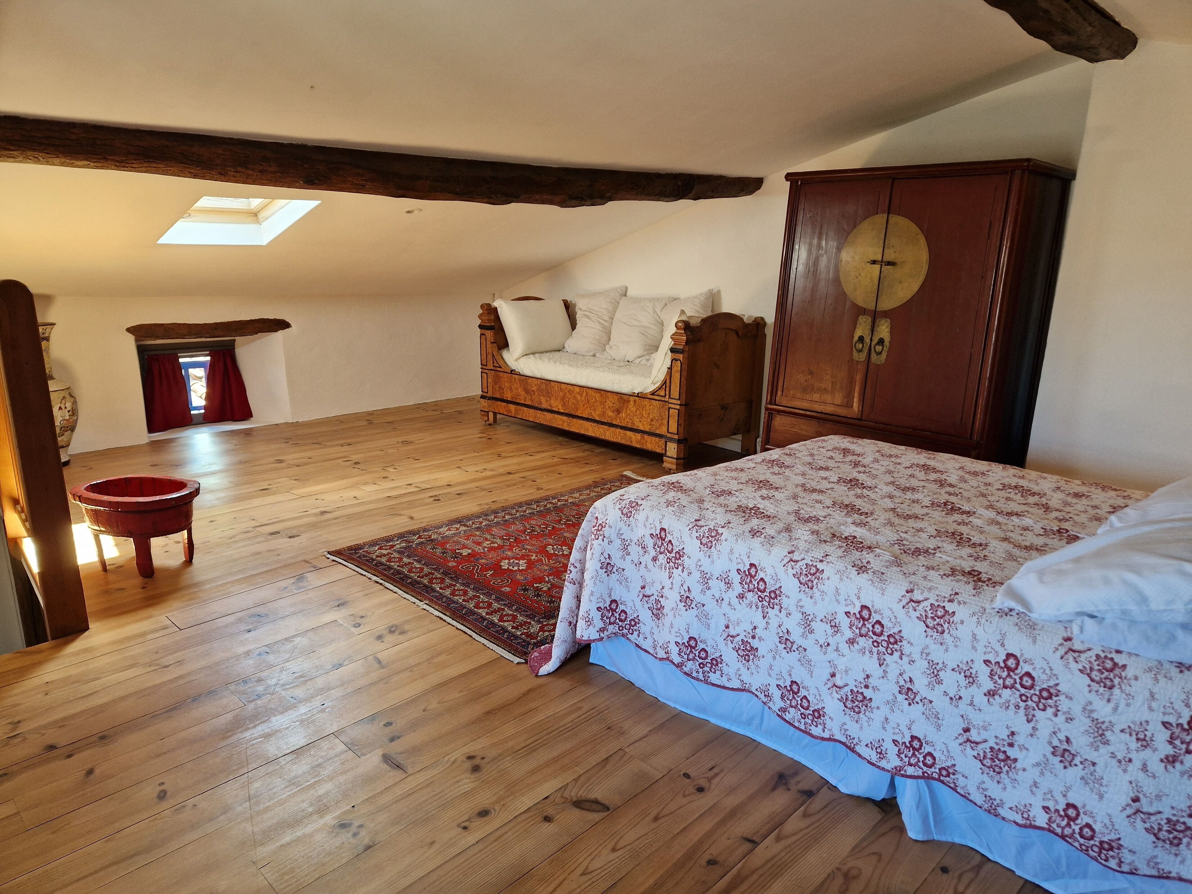 Un dormitorio con suelo de madera, una cama con colcha estampada, un armario de madera y un tragaluz.