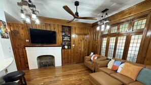 A cozy living room with a fireplace, wooden walls, a ceiling fan, and a large TV. - The Carriage House of Mount Vernon (Baltimore)