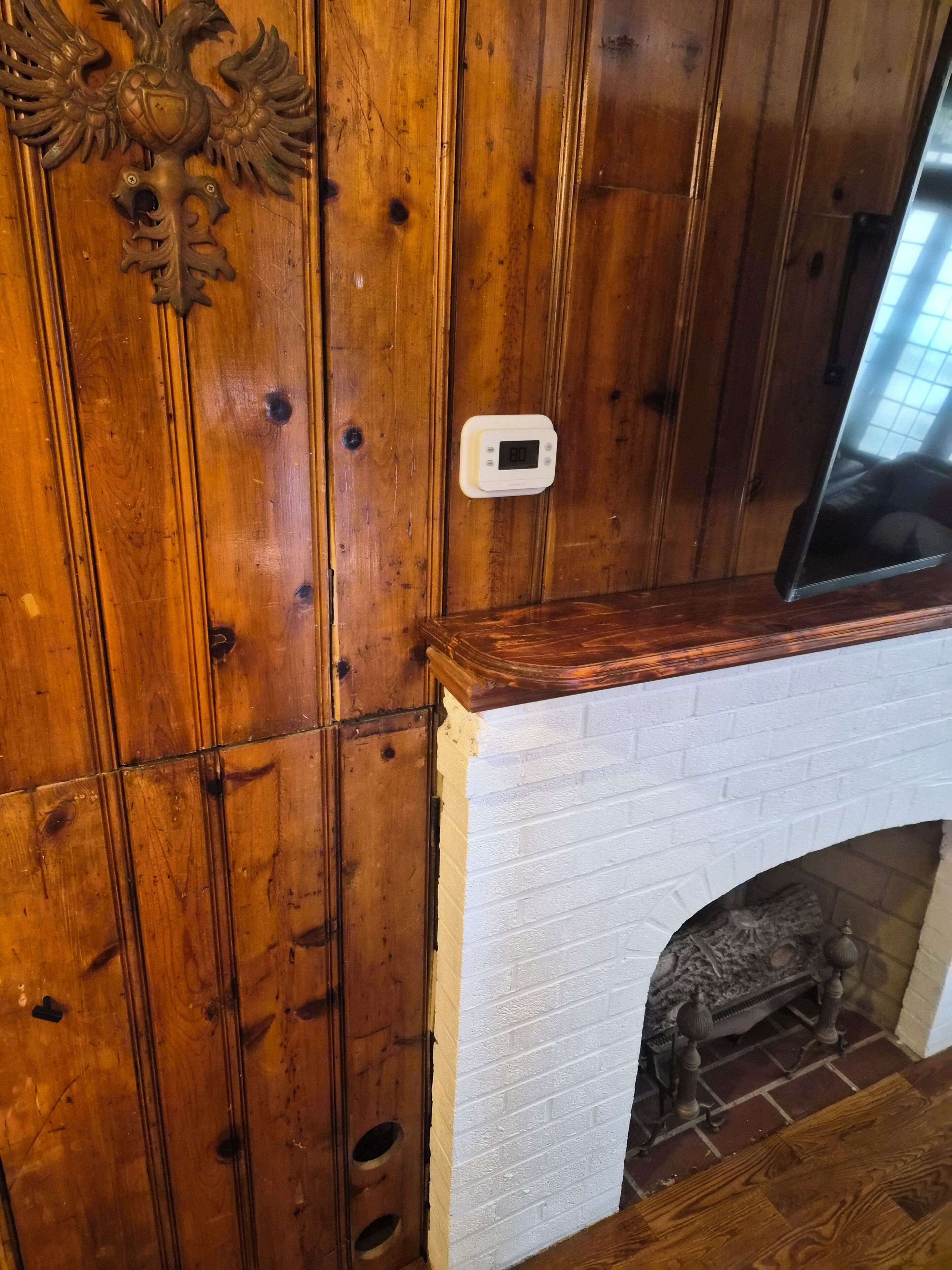 A wooden wall with a decorative emblem, a white brick fireplace, and a thermostat.