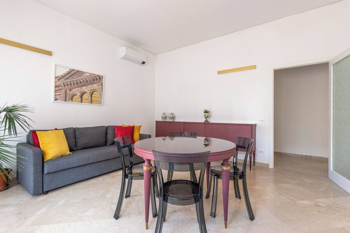 Una sala de estar moderna con un sofá gris, una mesa redonda de comedor con sillas, una planta en maceta y un aire acondicionado instalado en la pared.