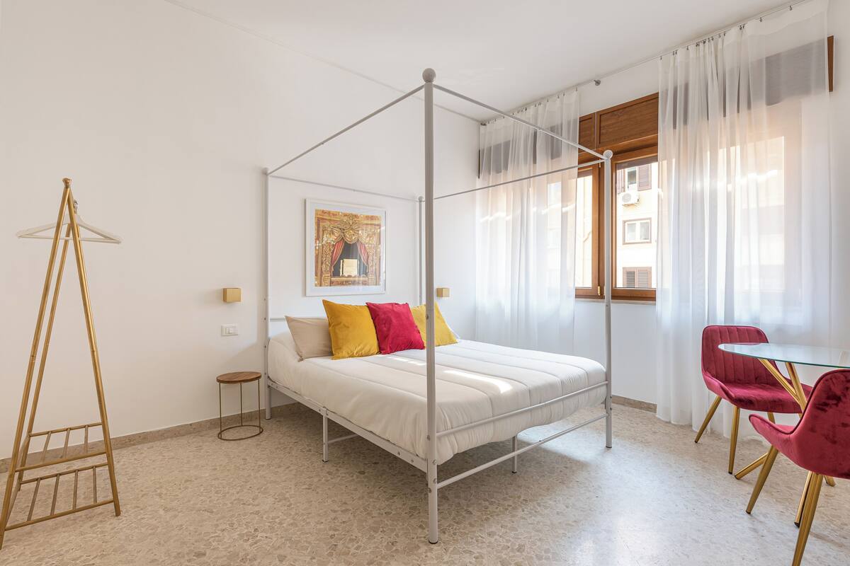 Una cama con dosel, ropa de cama blanca y almohadas coloridas, una mesita redonda con una silla, y un estante fijado a la pared.