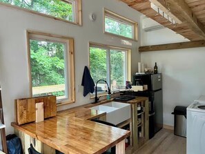 Una cocina con encimera de madera, un lavamanos, una cafetera y un refrigerador. Hay ventanas con vistas a árboles.