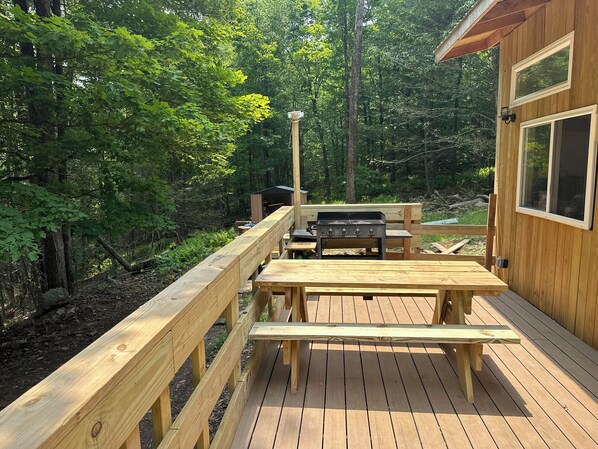 Una terraza de madera con una parrilla y una mesa de picnic, rodeada de un bosque.