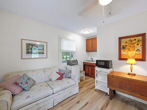 A furnished room with a sofa, a small kitchenette, a framed painting, and a window.