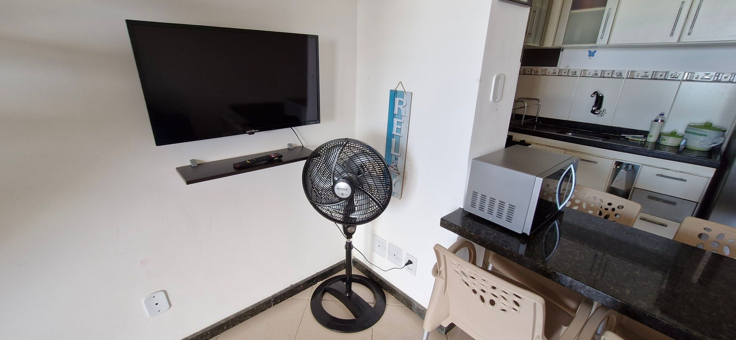 A compact kitchen with a flat-screen TV mounted above a shelf, a standing fan, and a microwave on the countertop.
