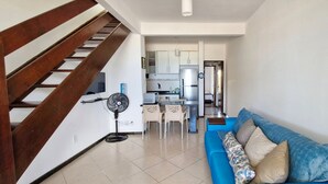 A modern living room with a blue sofa, a wooden staircase, a kitchen area, and a fan.