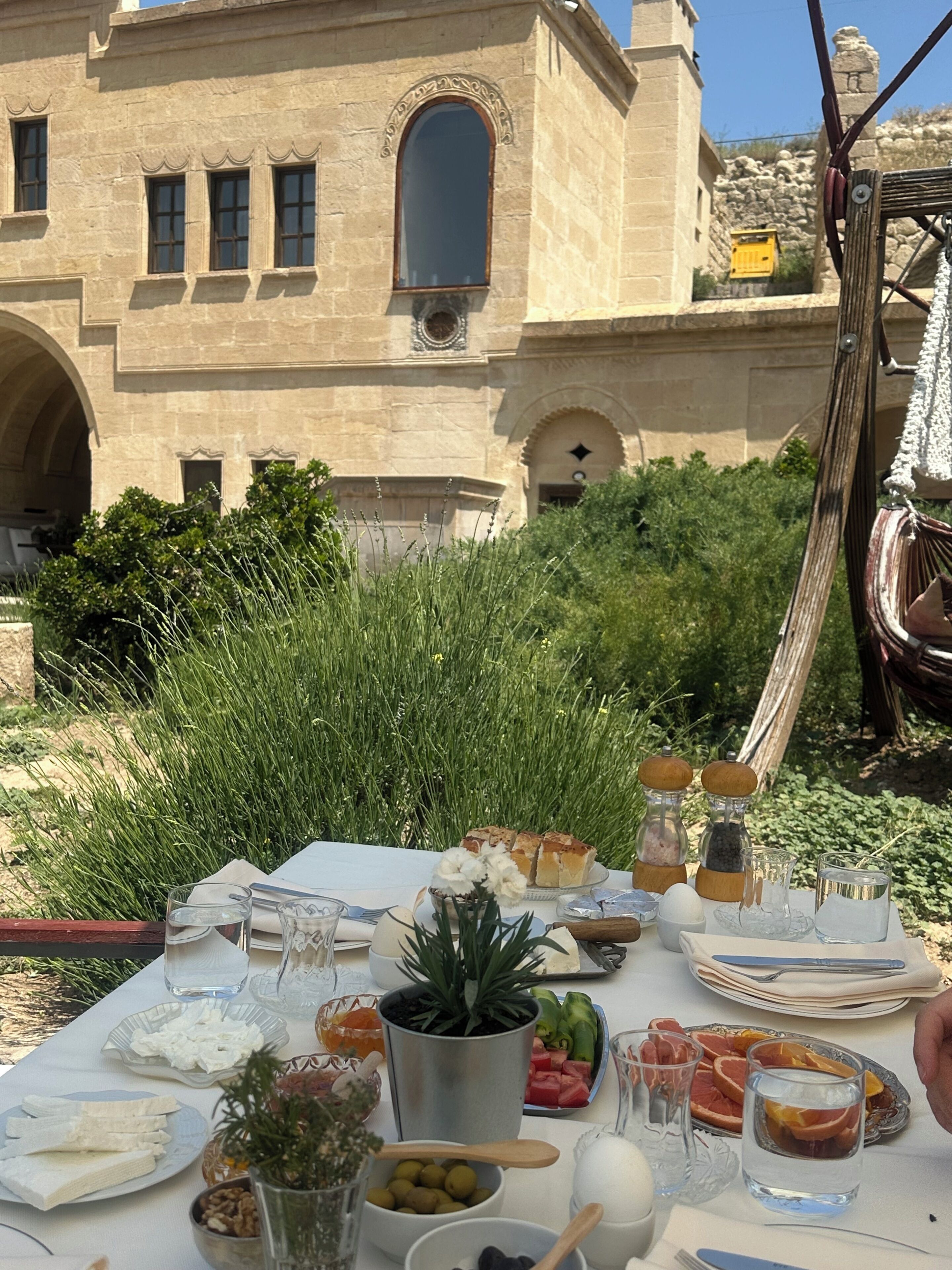 Mesa de comedor al aire libre con comida y bebidas, frente a un edificio con ventanas de arco y fachada de piedra.