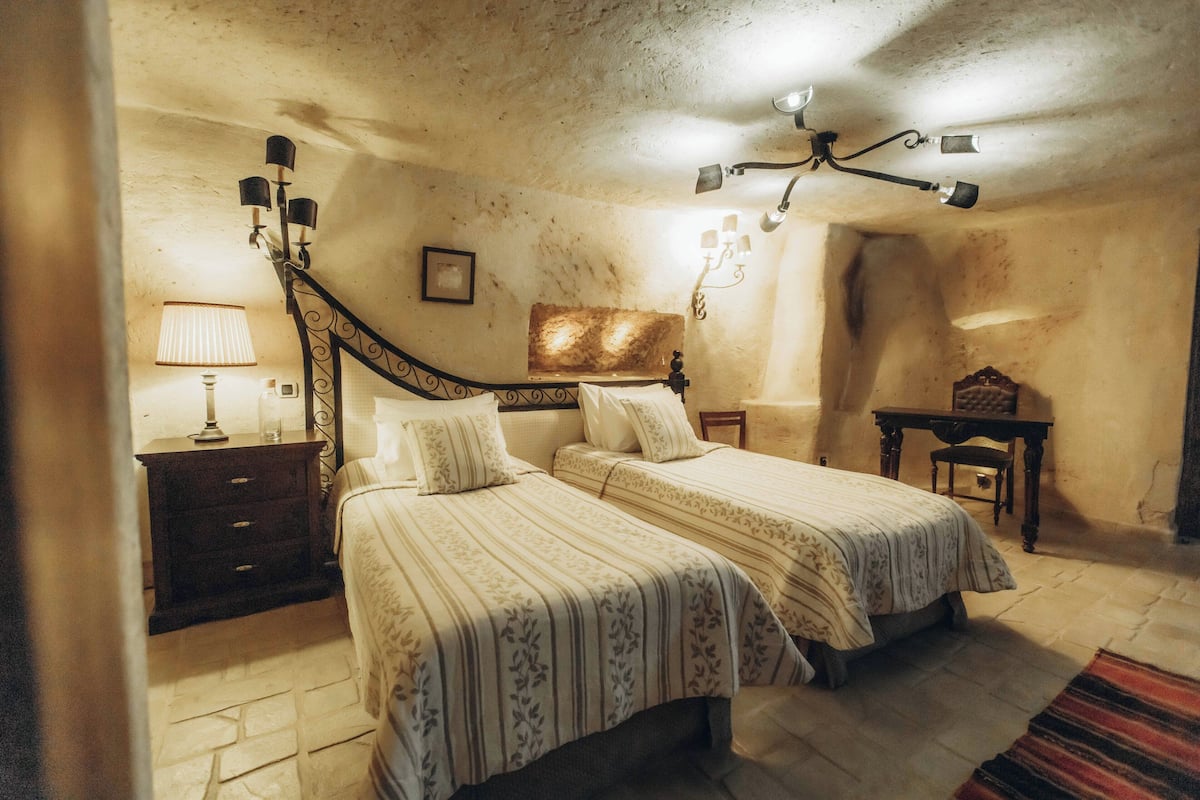 A bedroom with two beds, a nightstand, a lamp, a ceiling fan, and a small table with a chair.