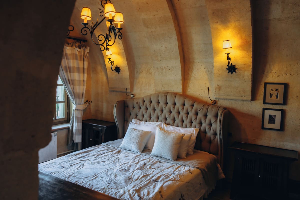 A bedroom with a tufted headboard, a large bed with white linens, a window with curtains, and wall-mounted lighting.