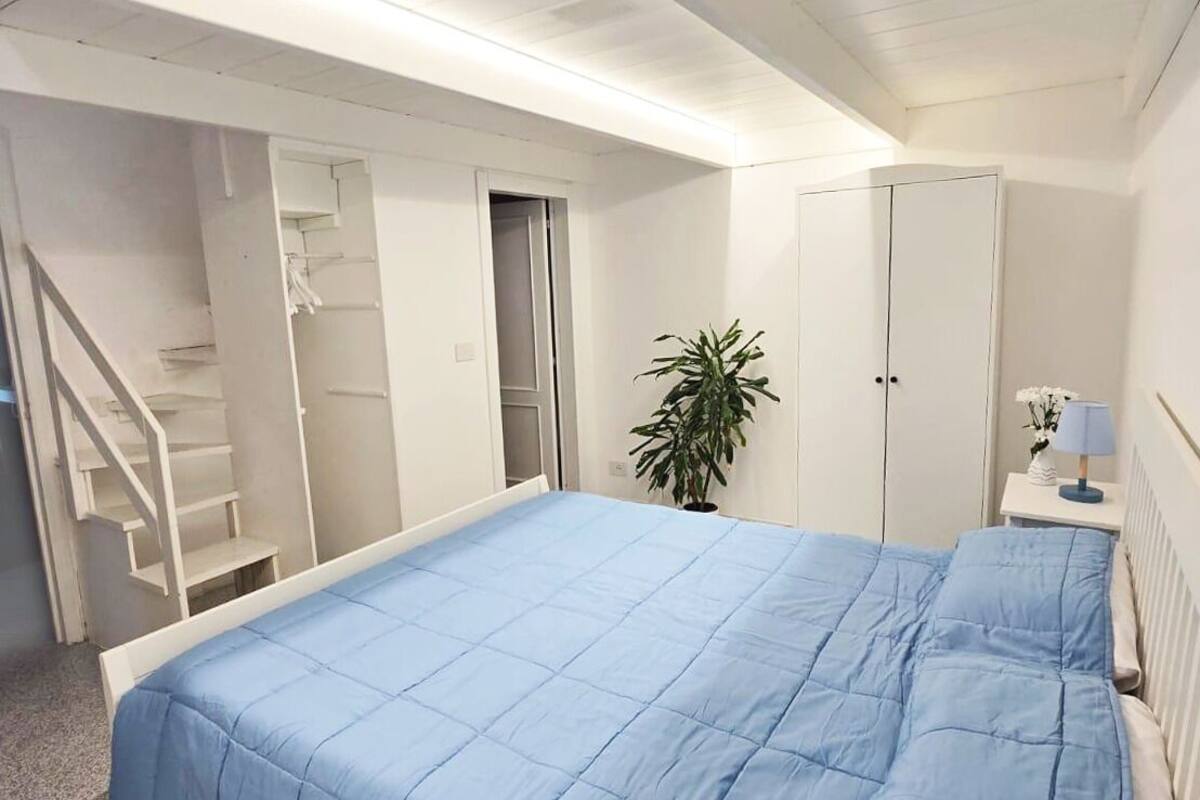 Un dormitorio con una cama grande, un armario blanco, una mesita de noche con lámpara y una escalera que lleva a un armario.