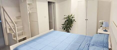 A bedroom with a large bed, a white wardrobe, a nightstand with a lamp, and a staircase leading to a closet.