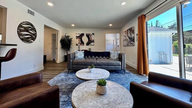 A modern living room with a grey sofa, two round coffee tables, and a large window with curtains. - 4BR · Pool · Fenced Yard · Garage · Tree-Lined Street (Carmichael)