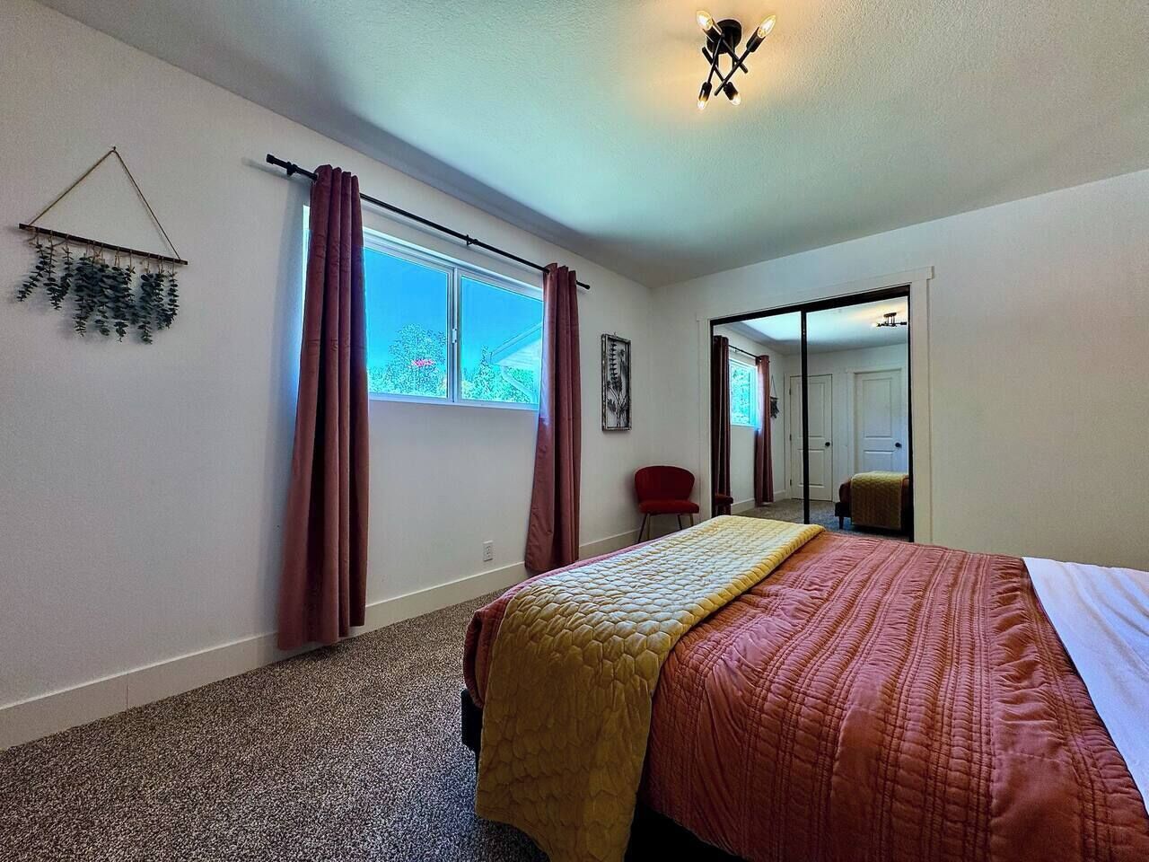 A bedroom with a bed, a window with curtains, a mirror, and a door leading to another room.