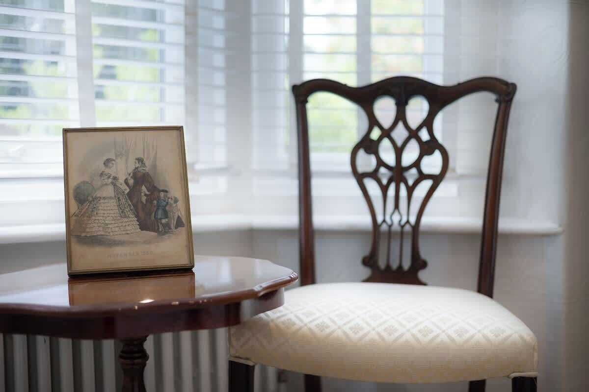 A framed picture on a table, a wooden chair with a patterned cushion, and a window with blinds.