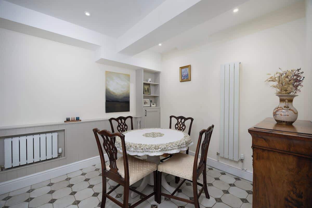 A dining area with a round table, six chairs, a radiator, a white tiled floor, a wooden cabinet, and a vase with dried flowers.