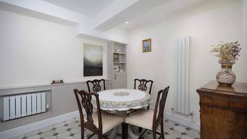 A dining area with a round table, six chairs, a radiator, a white tiled floor, a wooden cabinet, and a vase with dried flowers.