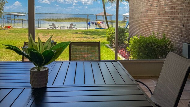 A patio with a table and chairs, a potted plant, and a view of a lake and distant buildings.