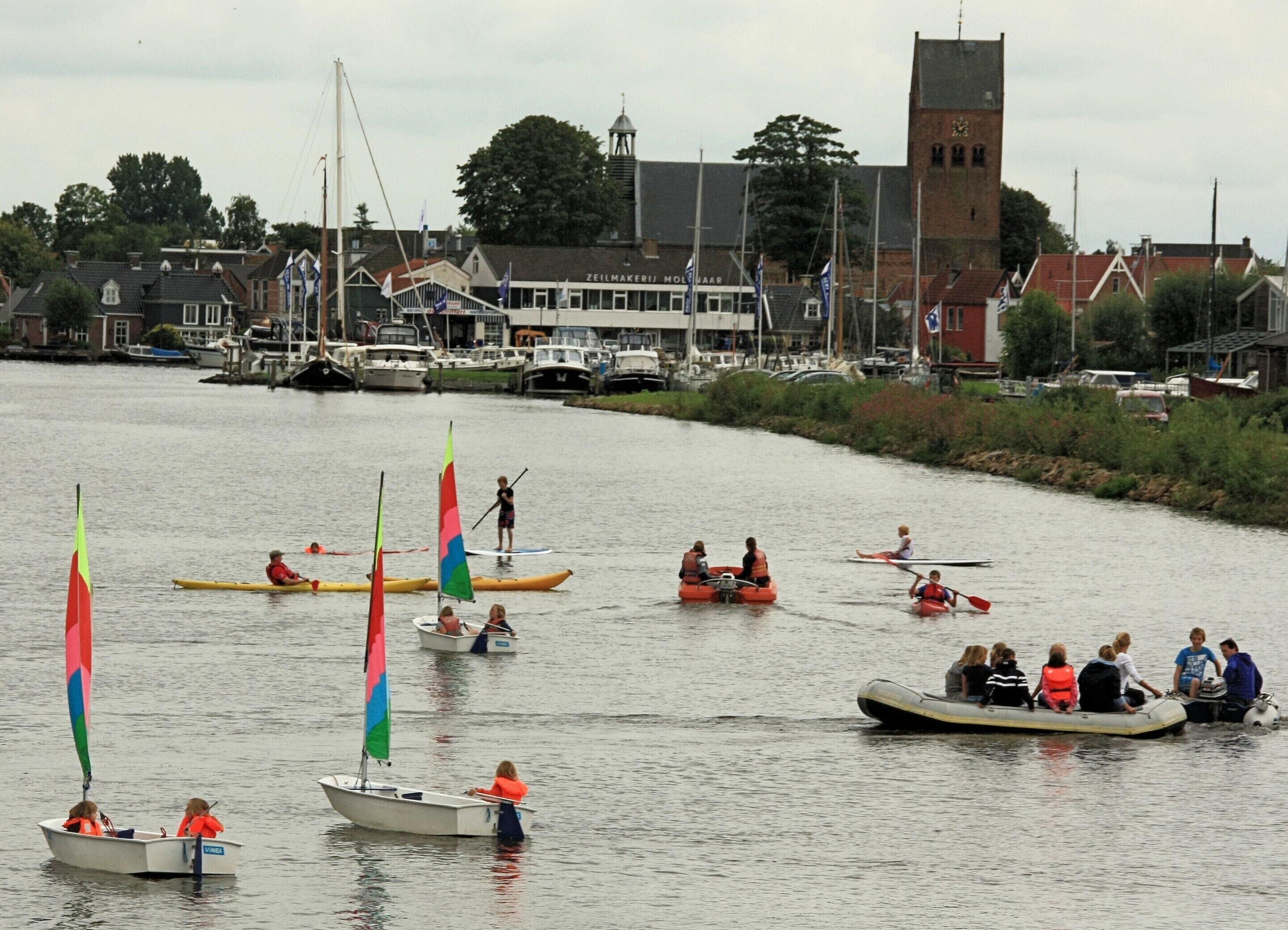A river with several small boats and canoes, some with colorful sails. There are buildings and a church tower in the background.
