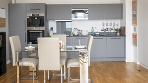 Een moderne keuken met grijze kastjes, roestvrijstalen apparatuur en een glazen eettafel met stoelen.