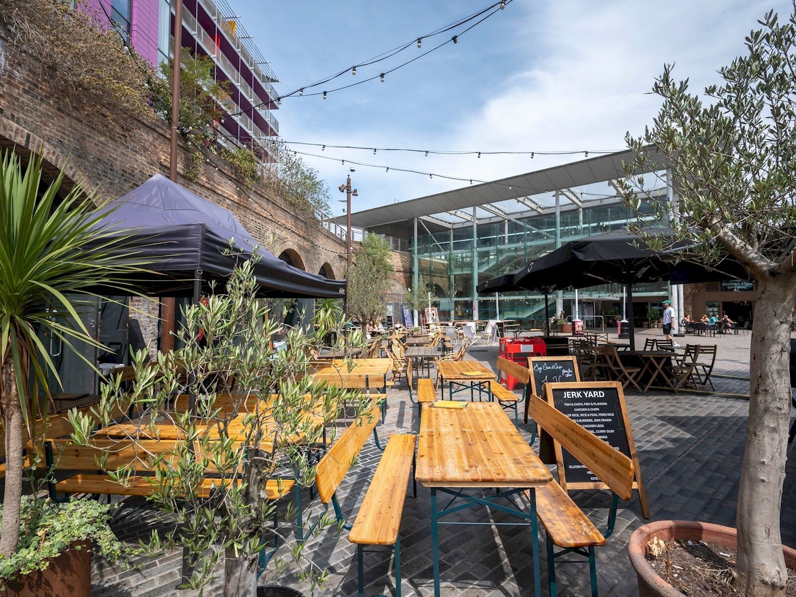 An outdoor seating area with wooden tables and benches, surrounded by potted plants and umbrellas.