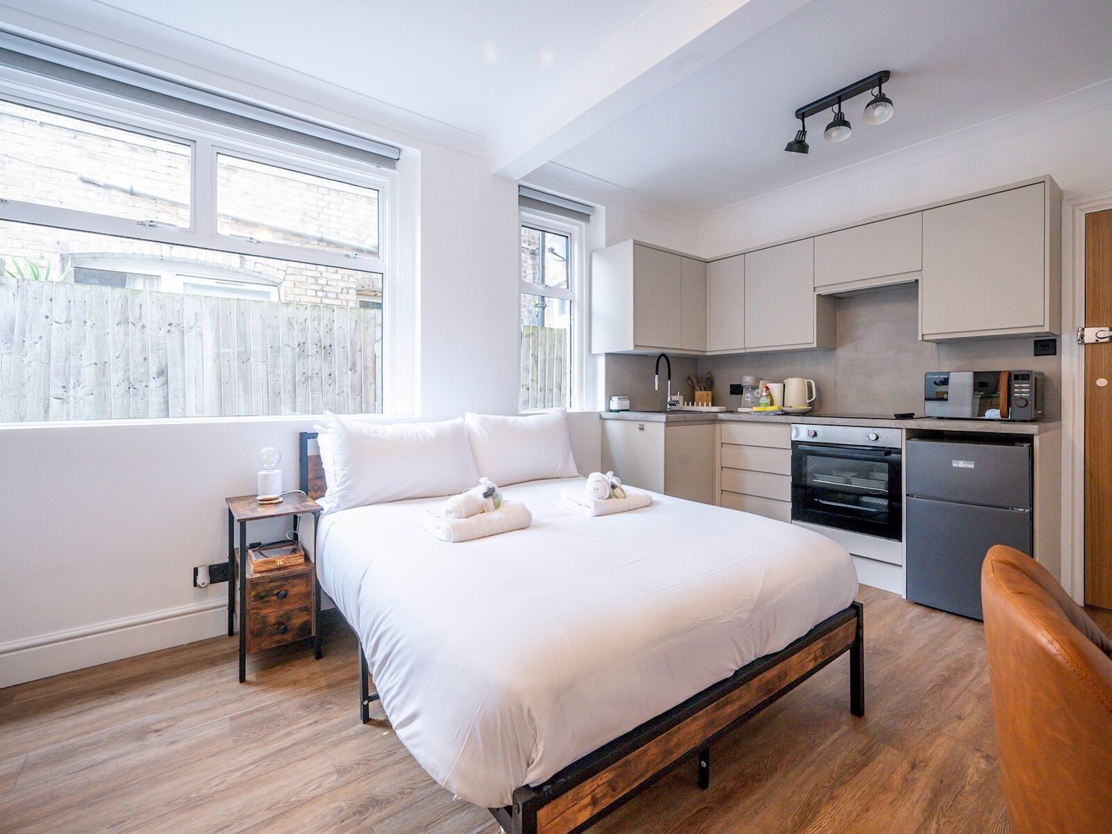 Een slaapkamer met een bed, nachtkastje en uitzicht op buiten. Grenzend daaraan een compacte kitchenette met fornuis, magnetron en kleine koelkast.