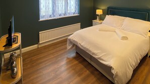 Une chambre avec un grand lit, une fenêtre ornée de rideaux légers, une table de chevet avec une lampe et un radiateur placé sous la fenêtre.