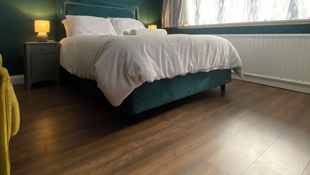 Une chambre à coucher avec un lit, une table de chevet, une lampe, une fenêtre avec des rideaux et un plancher en bois.