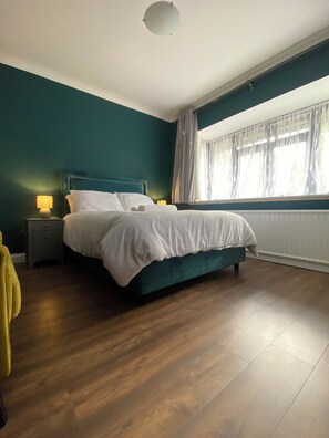 A bedroom with a bed, a nightstand, a lamp, a window with curtains, and a wooden floor.