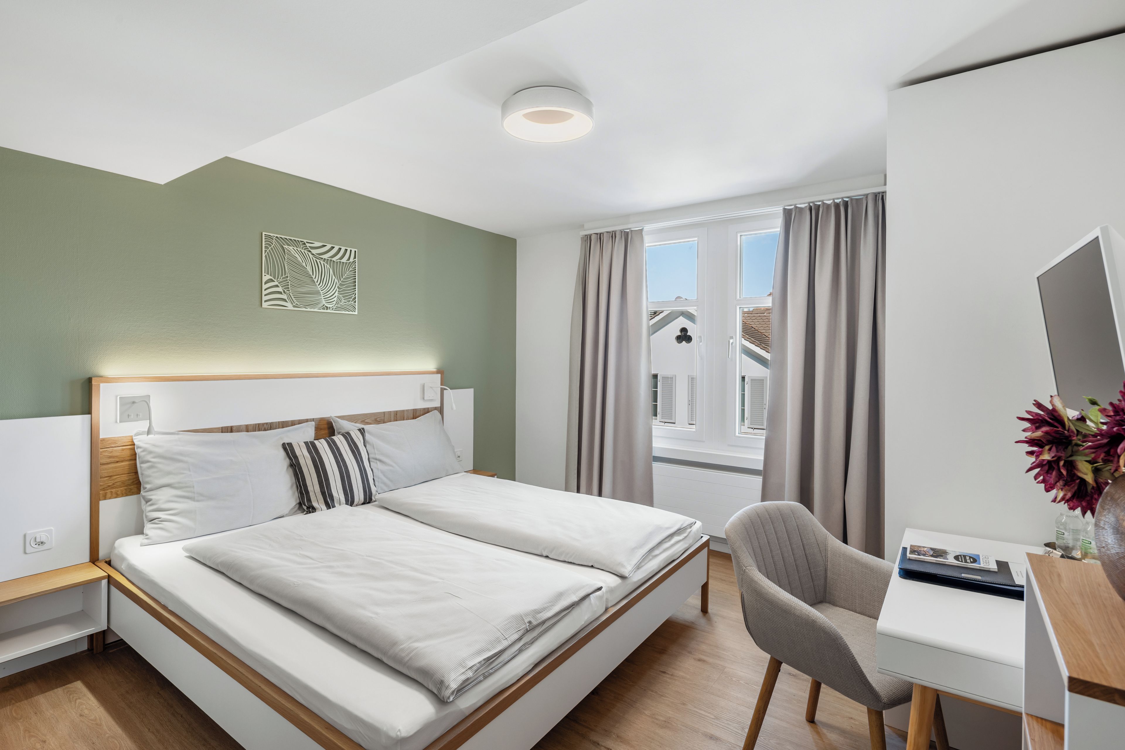 Quarto de hotel com cama, escrivaninha, cadeira, janela com cortinas e luz no teto.