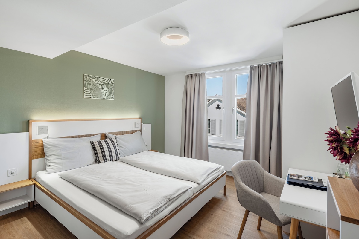 Une chambre d’hôtel avec un lit, un bureau, une chaise, une fenêtre avec des rideaux et un luminaire au plafond.