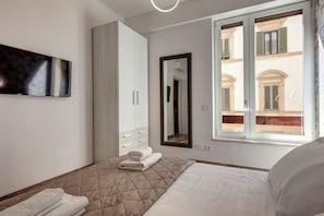Une chambre à coucher comprenant un lit, une armoire, un miroir et une fenêtre donnant sur des immeubles.