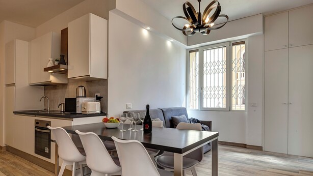 Una cocina moderna con armarios blancos, una mesa de comedor con cuatro sillas, una cafetera y una botella de vino.