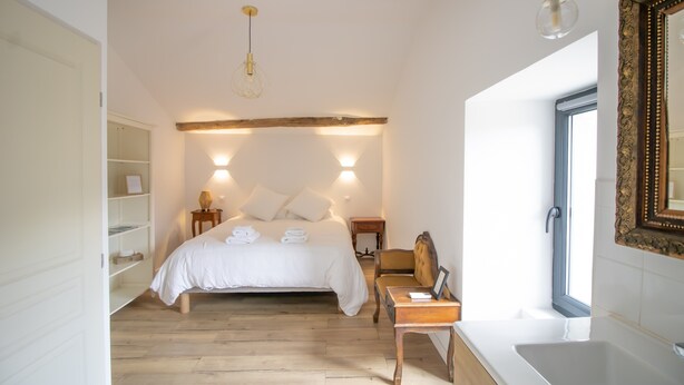 Een slaapkamer met een bed, nachtkastjes, een stoel, een bedlampje, een spiegel en een raam.
