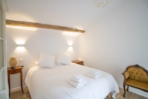 A neatly made bed with white linens and towels, a wooden nightstand with a lamp, and a vintage-style armchair.