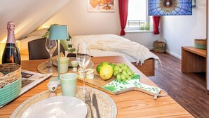 Une pièce chaleureuse avec une table à manger en bois, mise pour le repas, un lit recouvert d’une couverture blanche, un téléviseur fixé au mur et une fenêtre ornée de rideaux rouges.