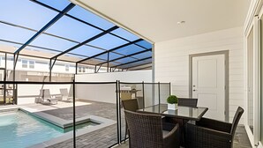 A covered outdoor patio with a glass table and chairs, a pool, and a door to the right. - Luxurious 5BD Space at Windsor Cay Resort (Clermont)