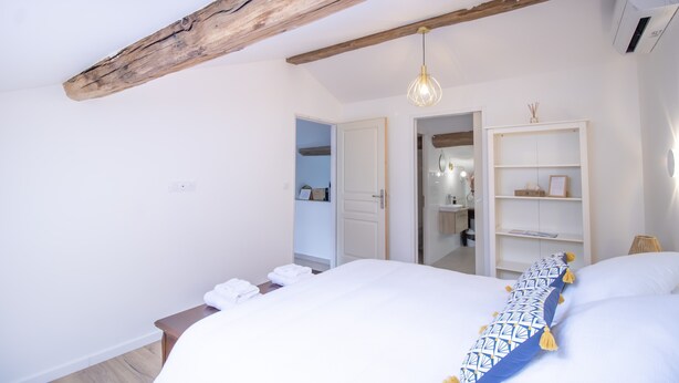 Une chambre à coucher avec un lit, une poutre en bois au plafond, un luminaire suspendu, une armoire et une porte donnant sur une autre pièce.