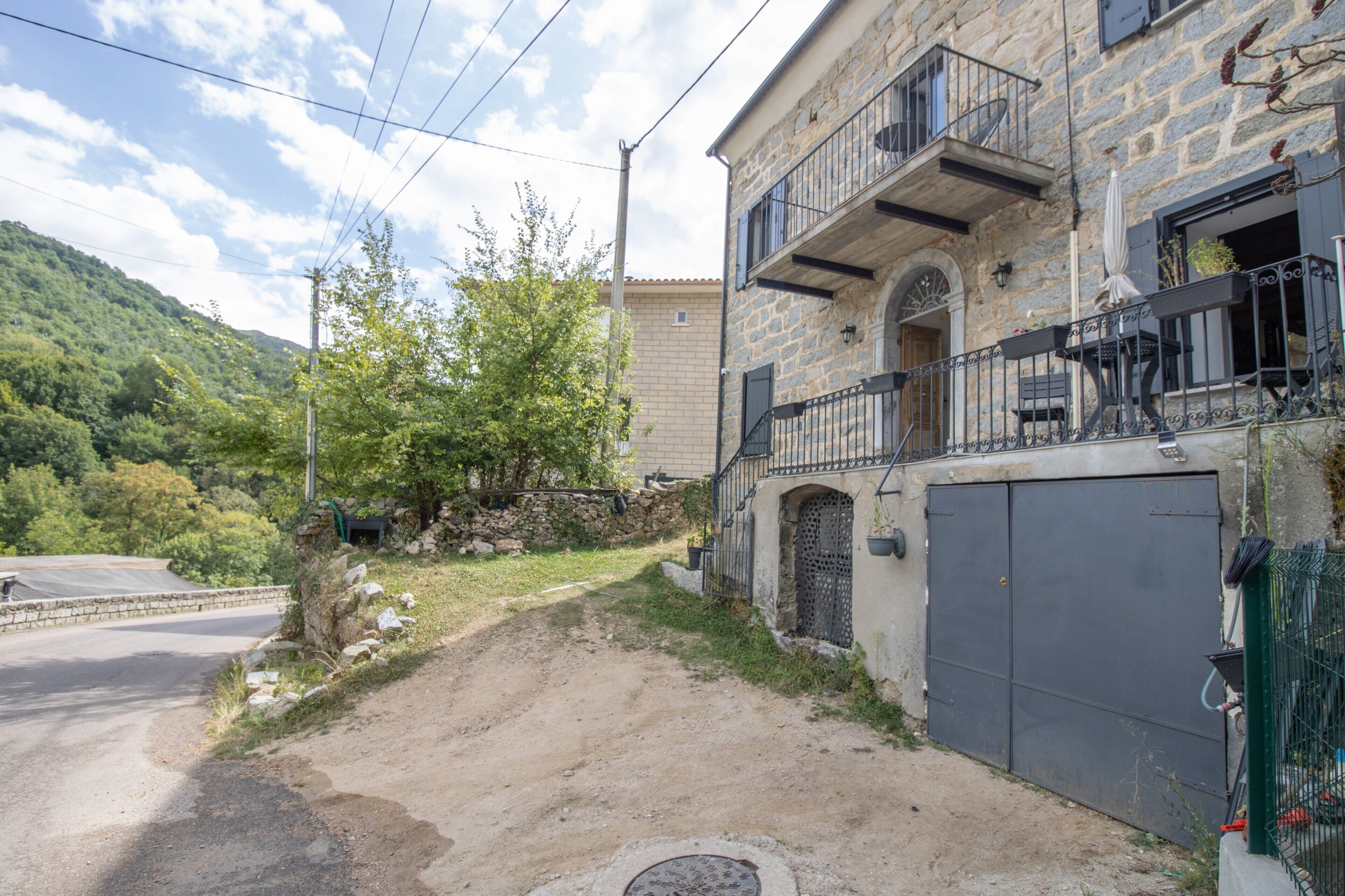 Ein Steinhaus mit Balkon, einer Garagenöffnung und einem Feldweg, der zur Straße führt.