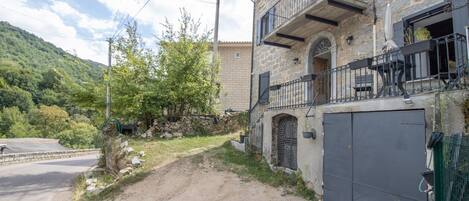Ein Steinhaus mit Balkon, einer Garagenöffnung und einem Feldweg, der zur Straße führt.